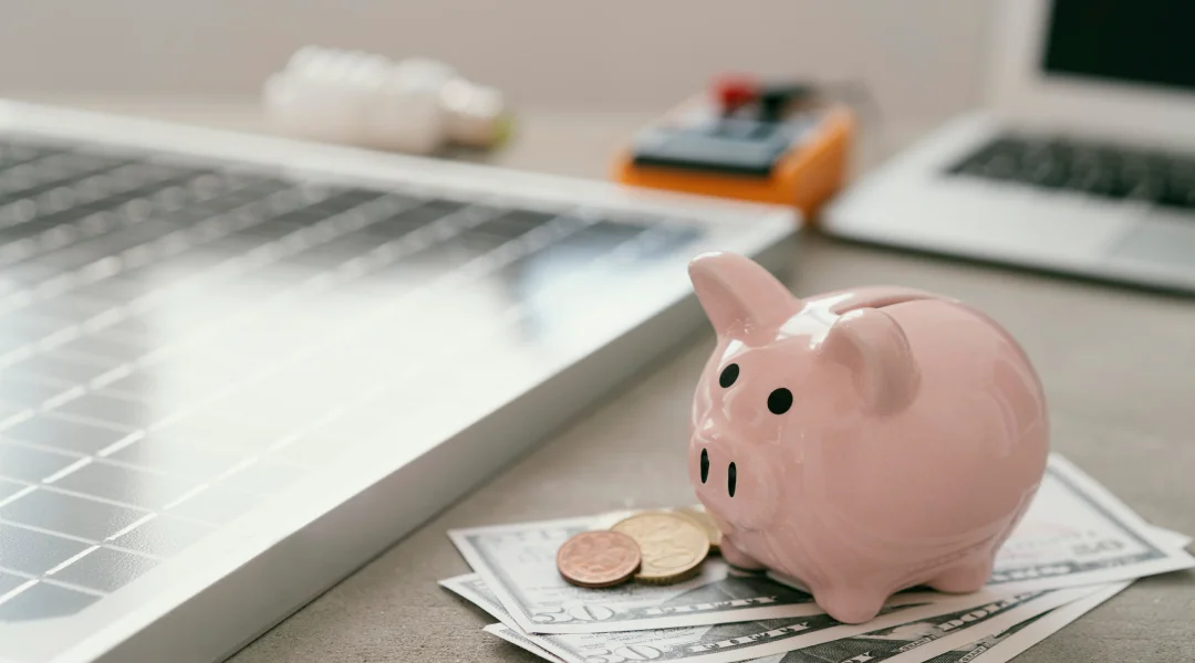 Piggy bank and cash on a desk with a laptop and solar panel, symbolizing strategies to get out of debt.