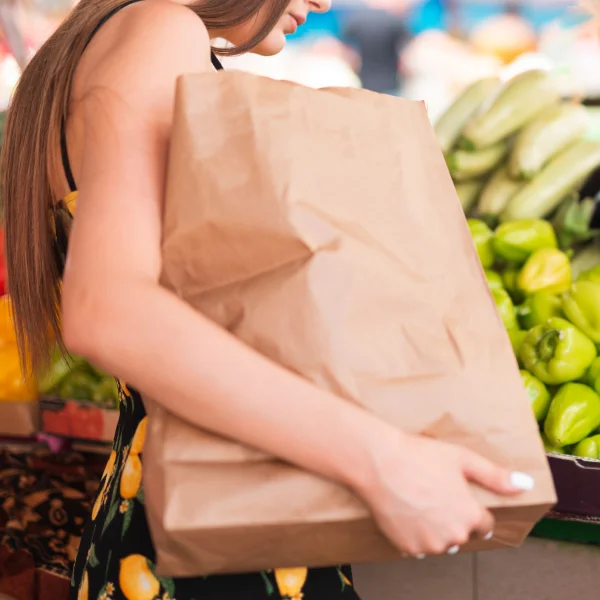 A woman with a brown paper bag stands in front of a produce stand, managing her budget while shopping for fresh fruits and vegetables.