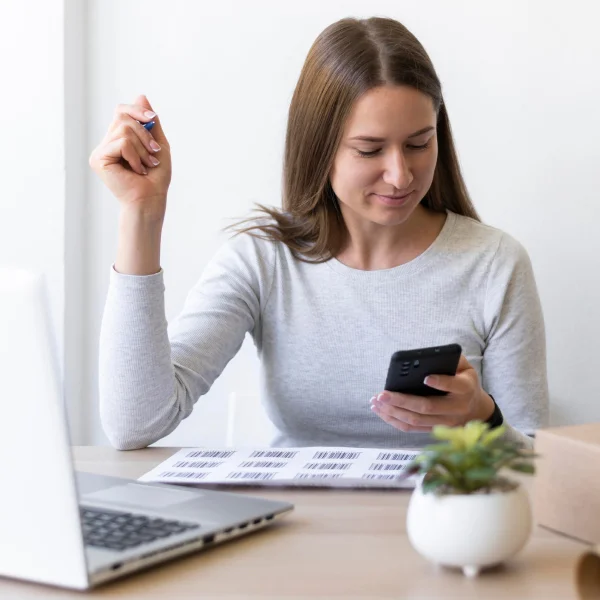 A woman sitting at a desk uses her cell phone, focused on managing her finances to get out of debt.