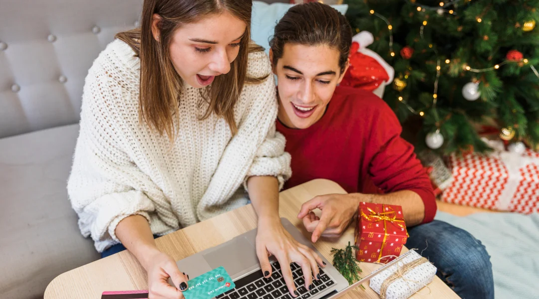 Un homme et une femme assis sur un canapé avec un ordinateur portable, devant un sapin de Noël décoré.
