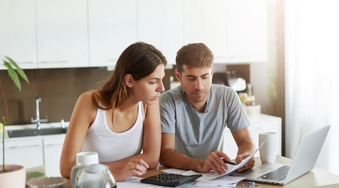 A man and woman sit at a table, focused on a laptop, discussing a short-term loan.