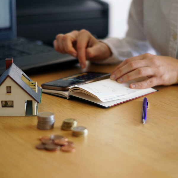 Person calculating expenses with a calculator next to a small house model, illustrating money management in an unstable economic context.